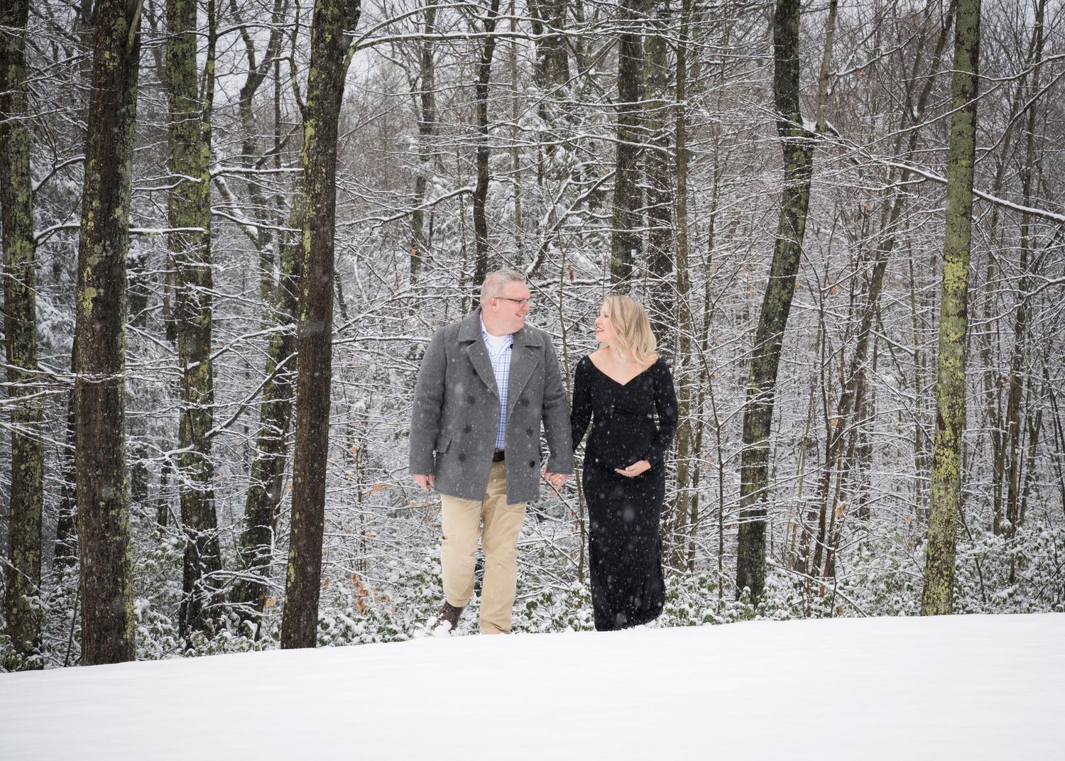 A couple standing together in a snowy winter forest with snow-covered trees creating a romantic woodland setting.