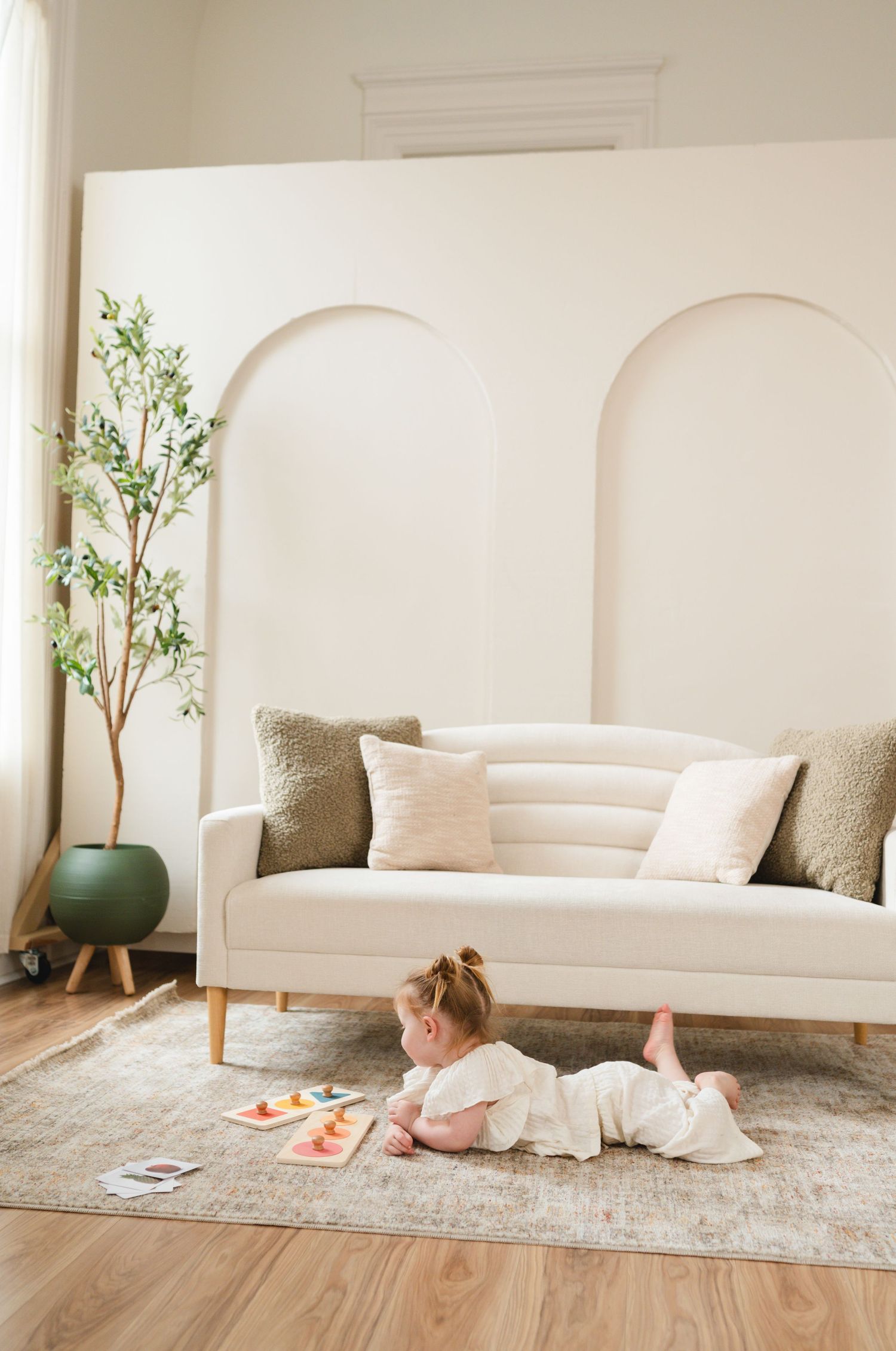 Child plays on beige area rug near white modern sofa and potted plant in minimalist living room with arched wall details.
