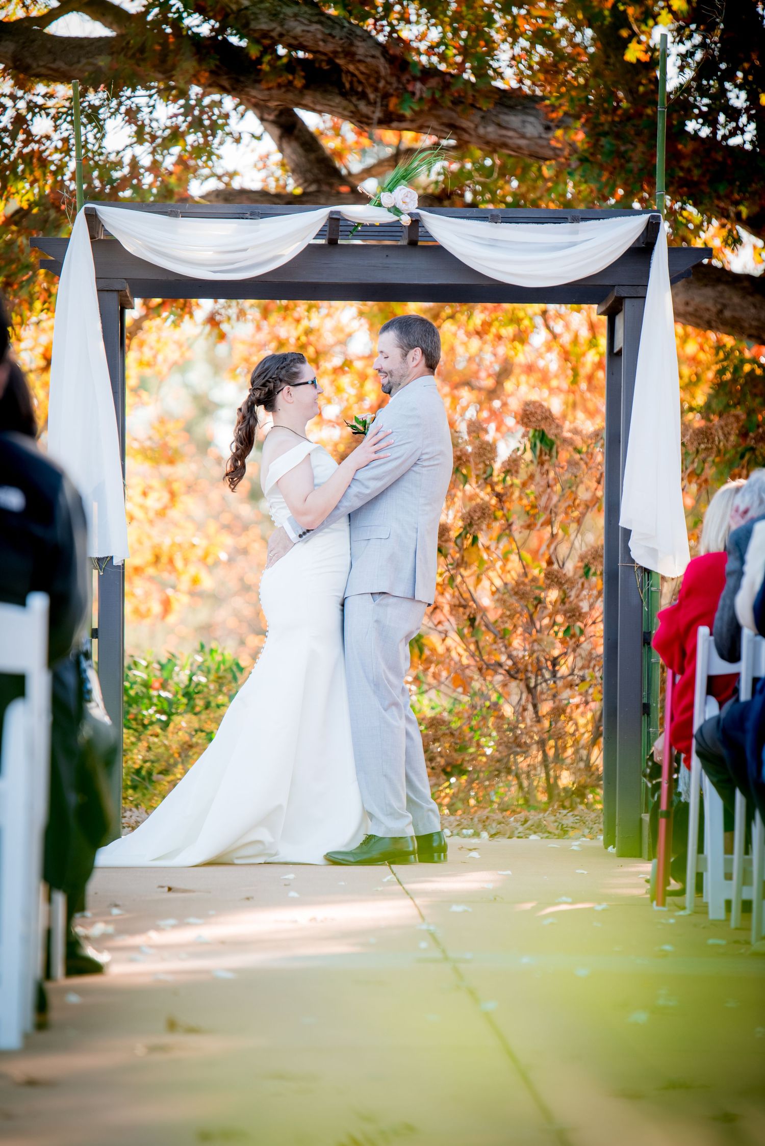 A couple shares their first kiss under a white draped arch during an autumn outdoor wedding ceremony.