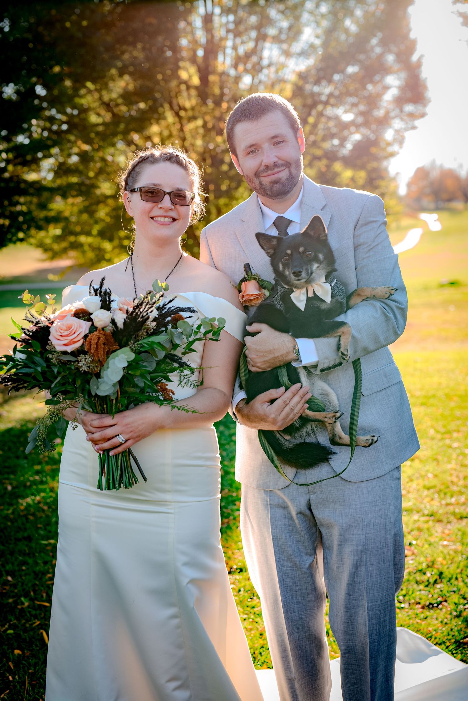 A couple in wedding attire stand outdoors holding floral bouquets and pets on a sunny autumn day.