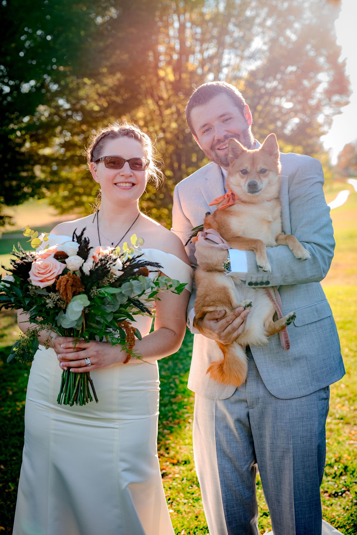 A couple in wedding attire poses outdoors at sunset with their small tan dog while holding a floral bouquet.