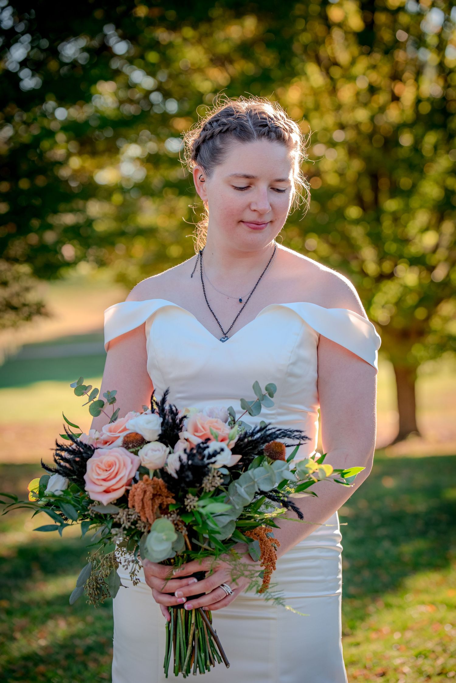 A bride in an off-shoulder white dress smiles while holding a beautiful peach and green bouquet in golden sunlight.