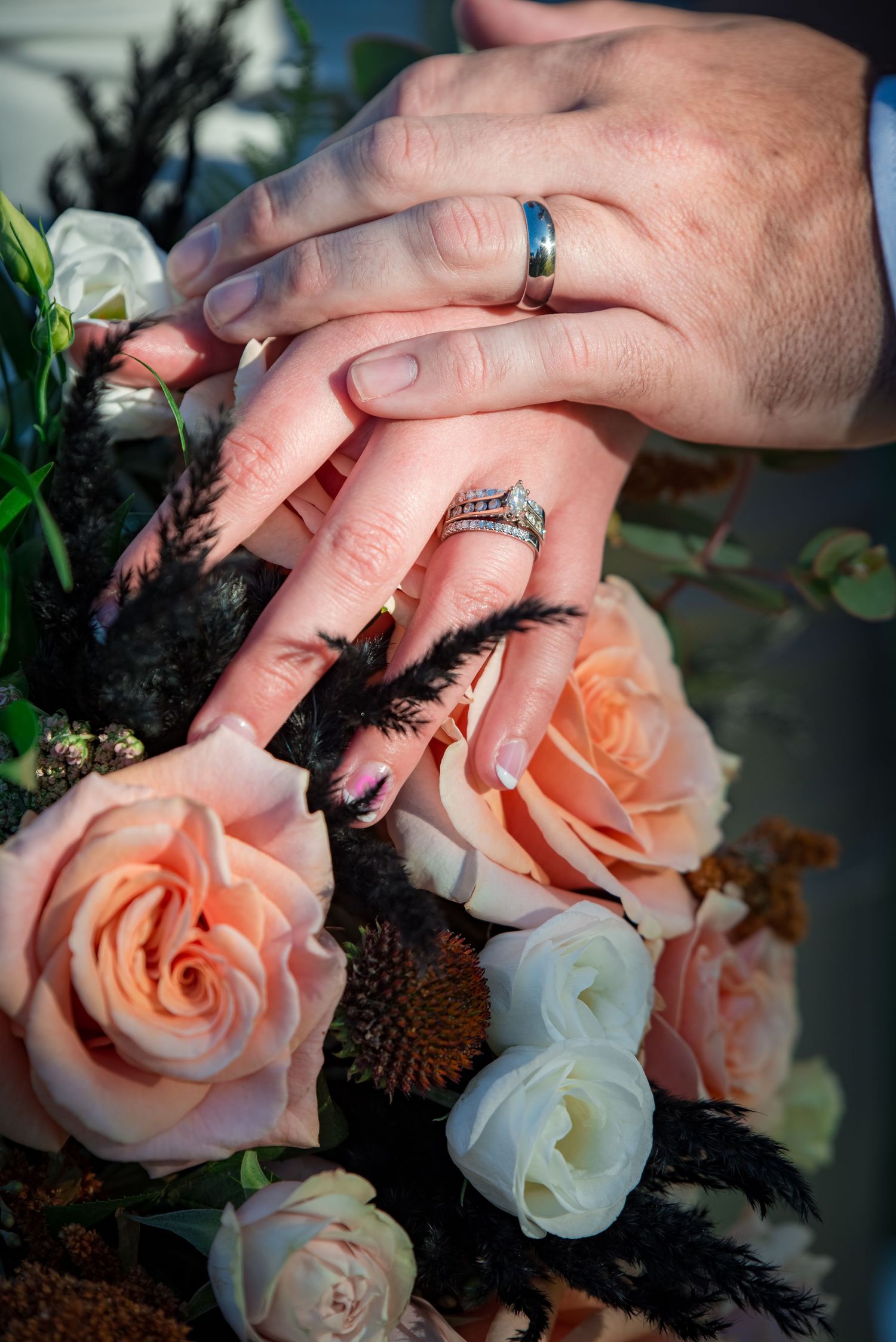 Wedding rings displayed on hands with peach and white roses and black feather accents.