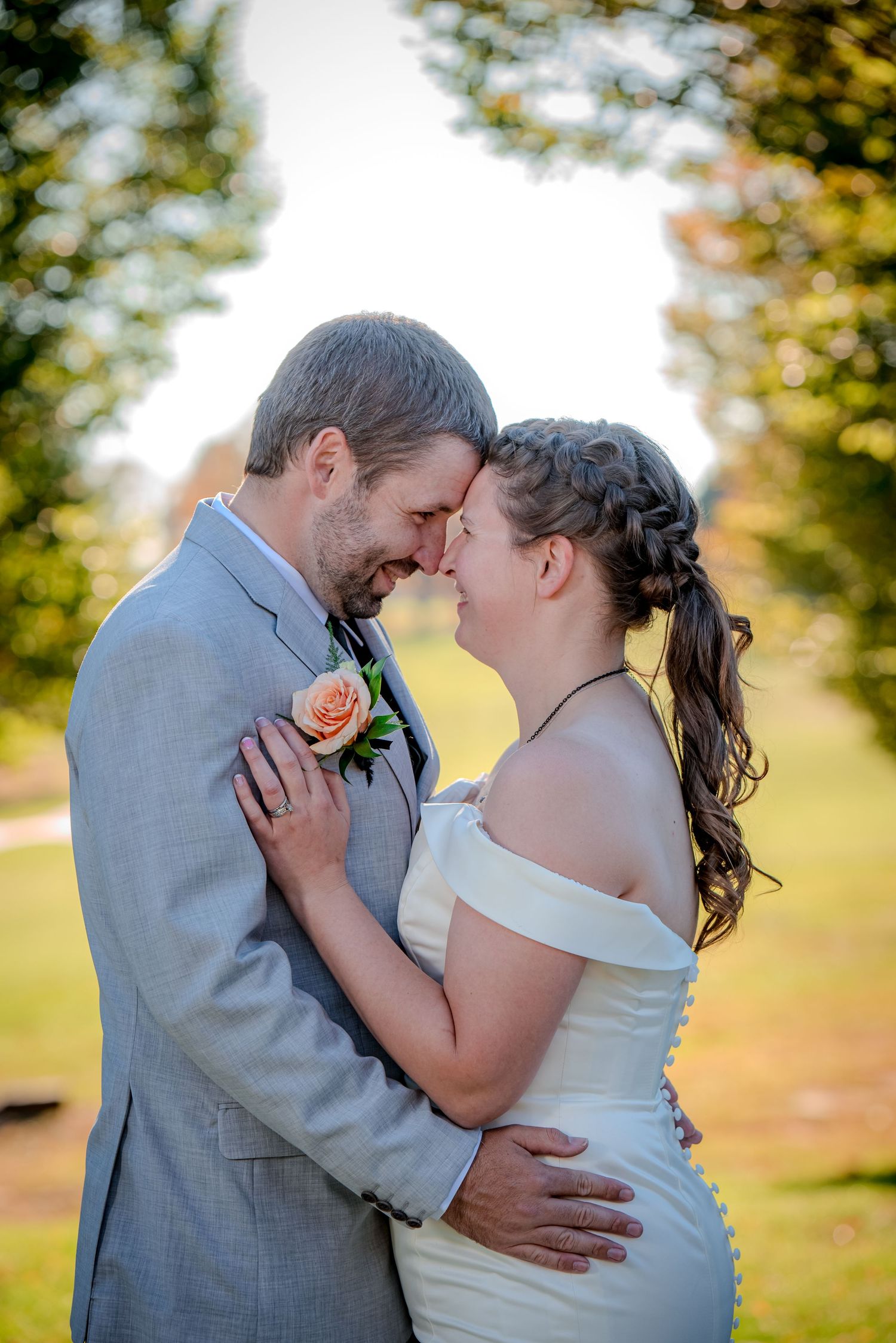 A tender romantic moment between newlyweds embracing outdoors during golden hour with a peach rose boutonniere.