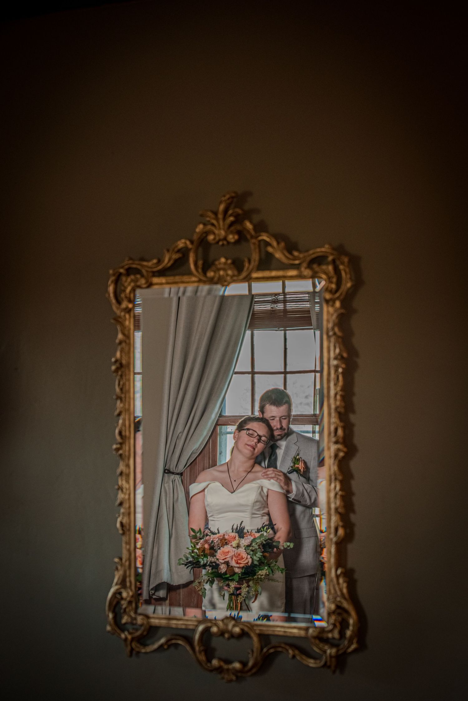 A decorative gold baroque mirror reflects a romantic wedding moment with a bouquet of pink and peach flowers.