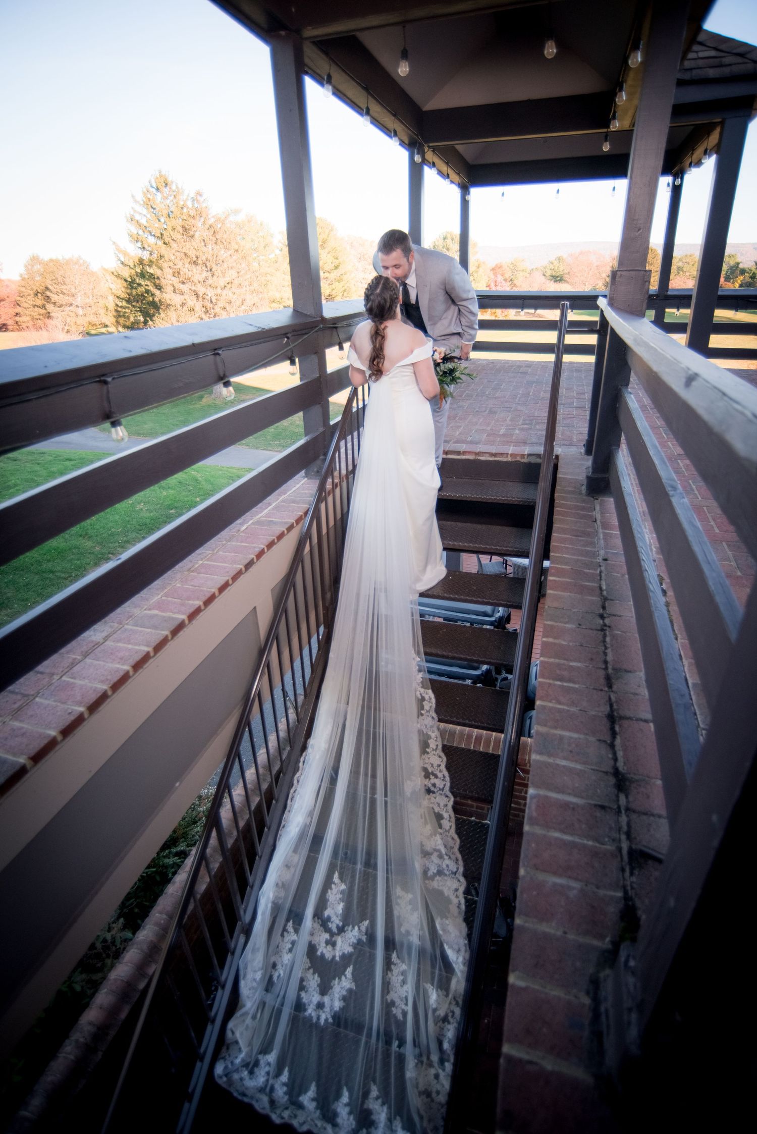 A long lace wedding veil dramatically trails down wooden stairs as couple embraces on covered porch.