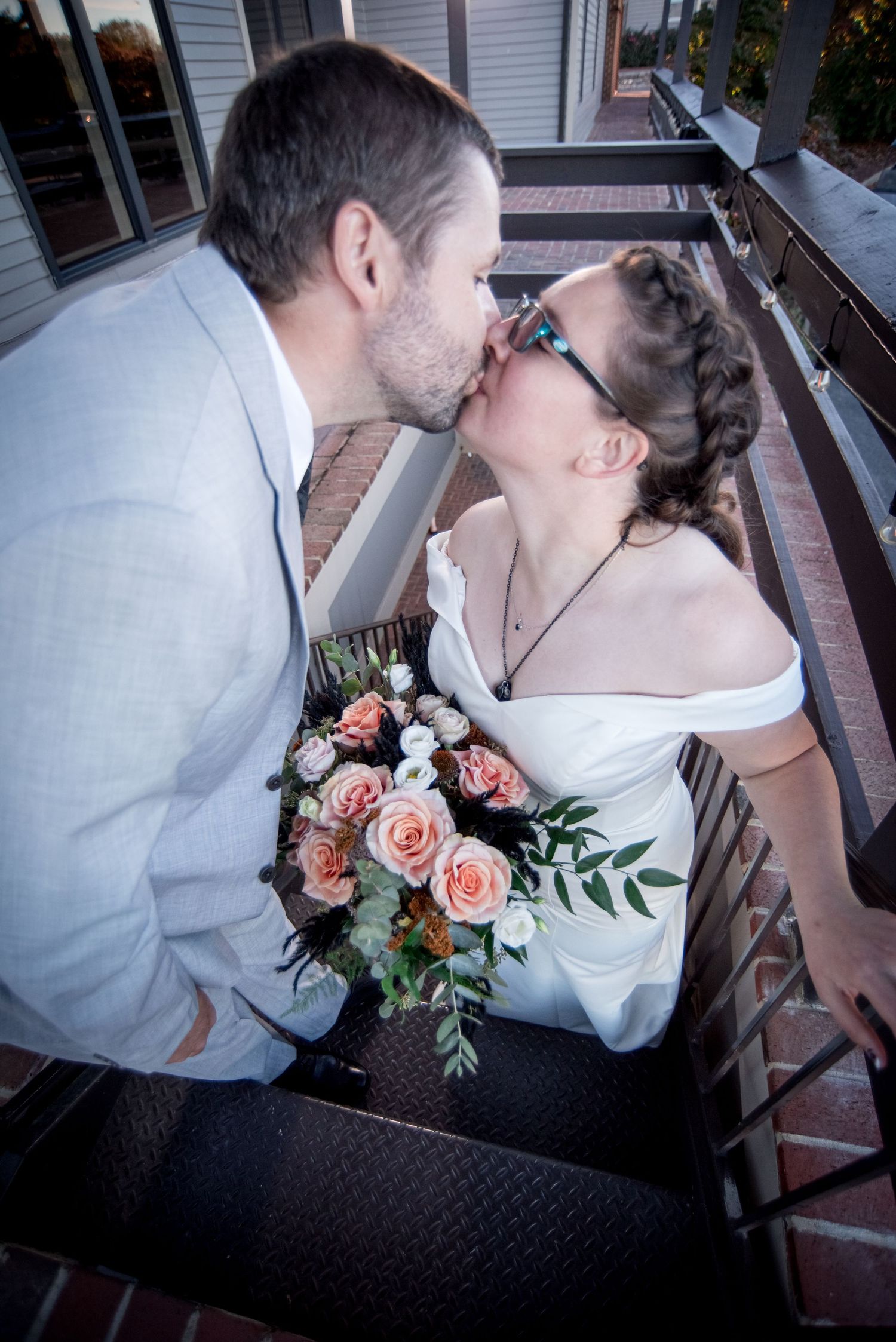 A wedding couple shares a romantic kiss on porch stairs while holding a peach rose bouquet.