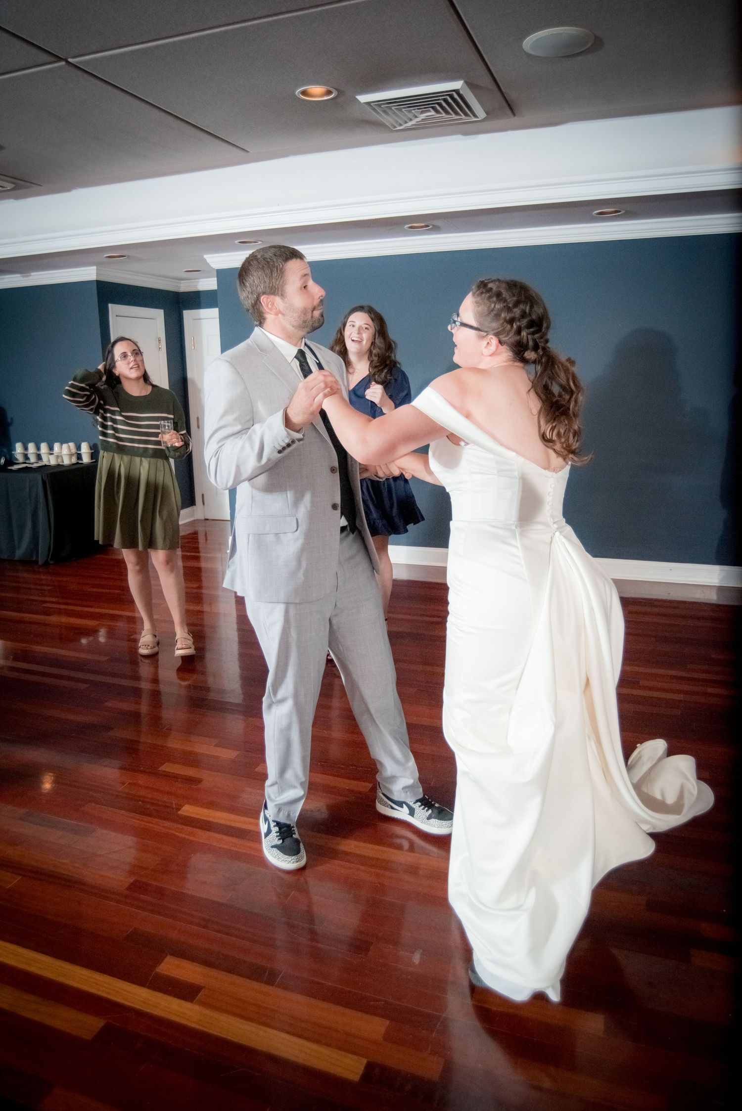 A couple dances at their wedding reception on a hardwood floor with a guest looking on in the background.