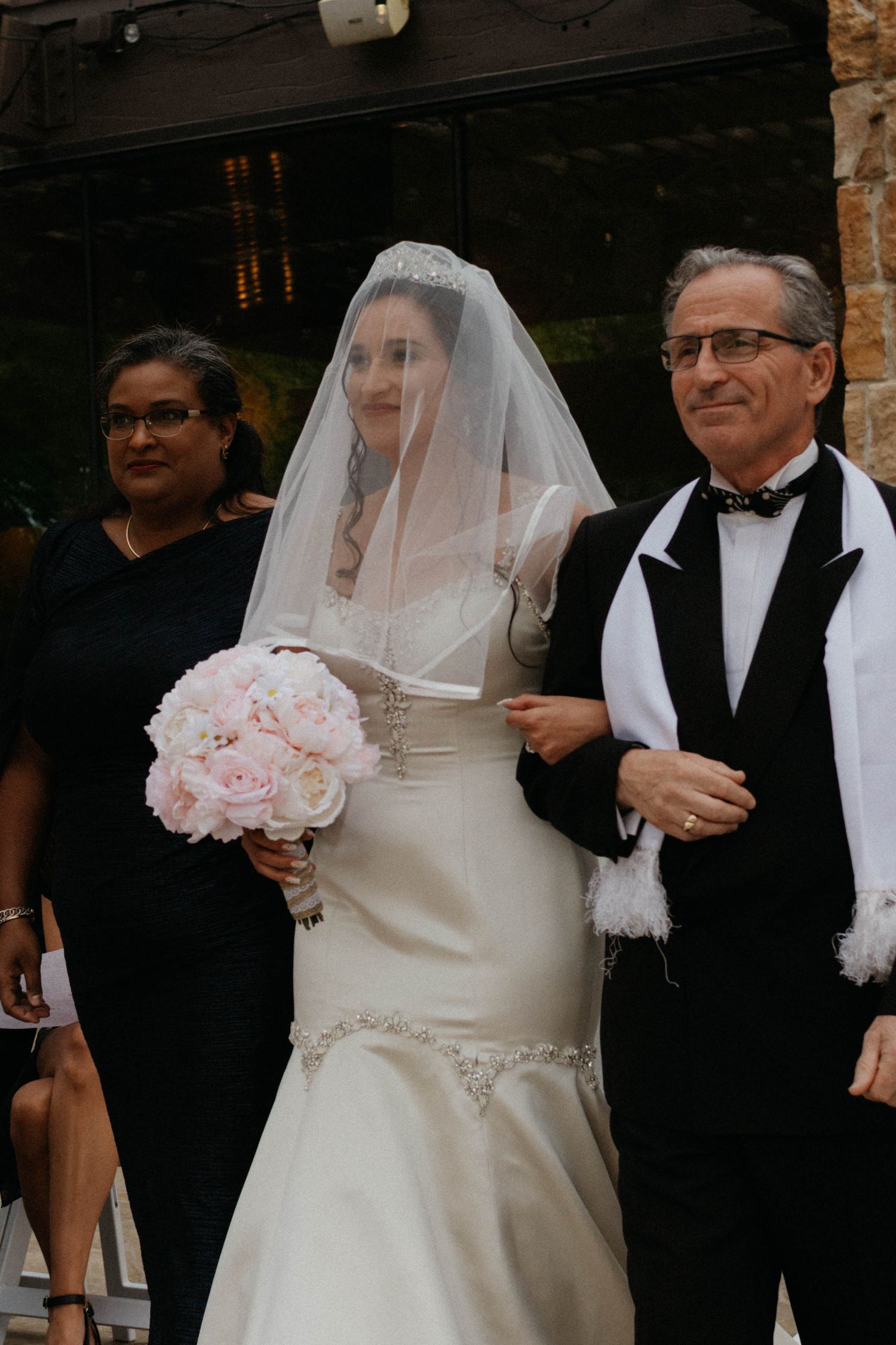 A bride in a white mermaid wedding gown and veil holds a pink rose bouquet while being escorted into a venue. Colorado Wedding photographer