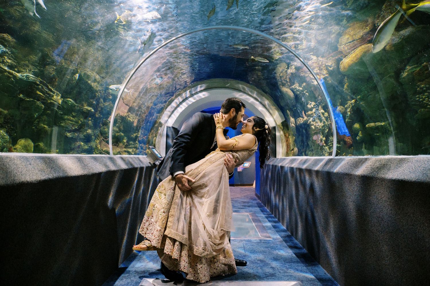 A couple embraces in a romantic moment under a curved aquarium tunnel with fish swimming overhead.