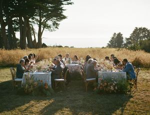 Outdoor wedding reception dinner in golden field with guests seated at floral-decorated tables surrounded by tall trees.