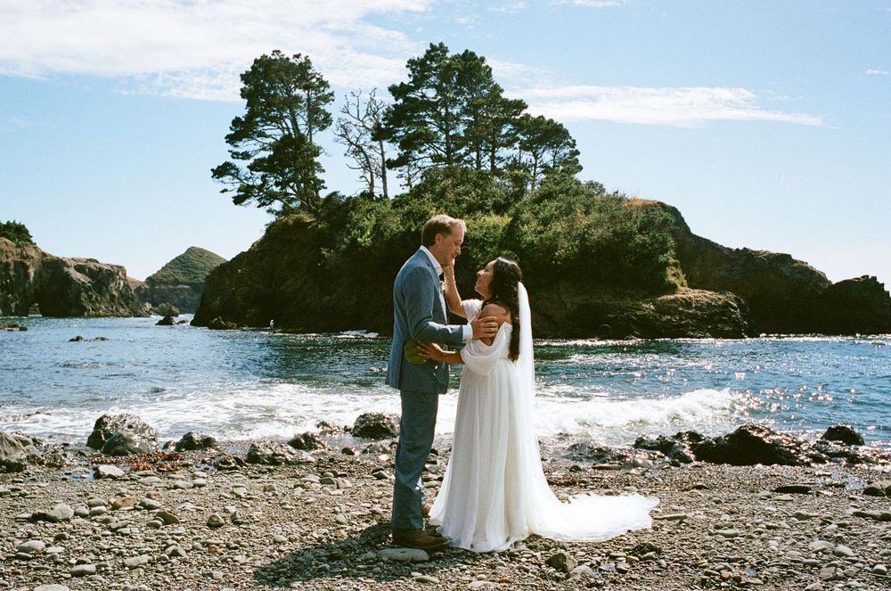 Couple exchanging vows on a rocky beach with ocean waves and tree-covered rock formations in background.