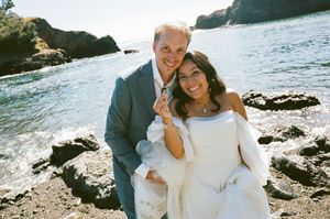 Happy couple in wedding attire smiling on rocky coastal beach with ocean waves and cliffs in background.