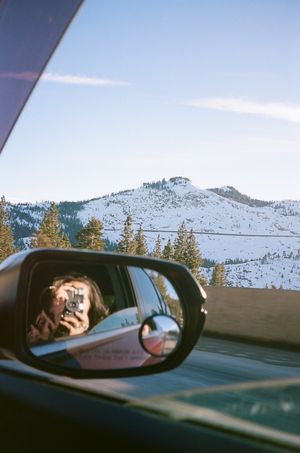 Snow-capped mountain peaks and evergreen trees are reflected in a car's side mirror on a winter day.