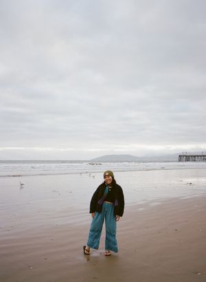 A person in a black jacket and wide-leg jeans stands on a misty beach with a pier visible in the distance.