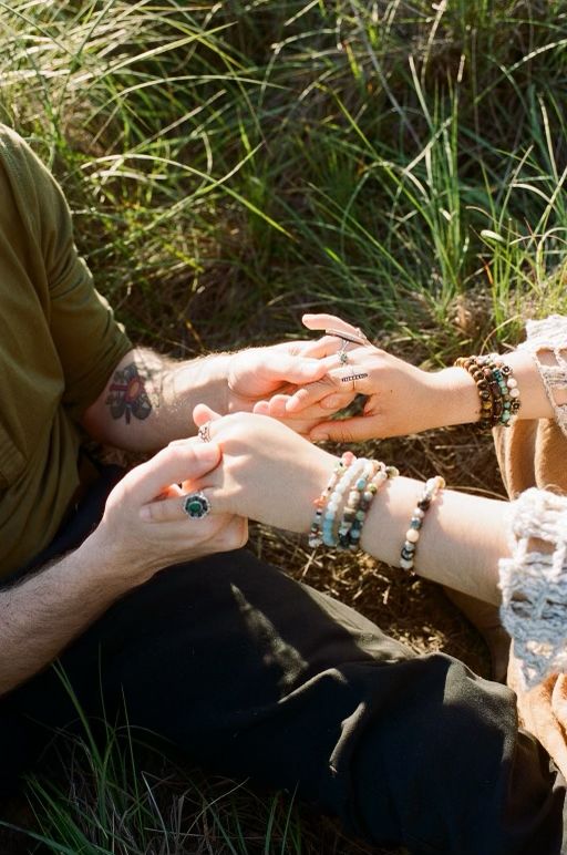 Multiple hands wearing bohemian beaded bracelets and rings touch in sunlit grass during a peaceful outdoor moment.