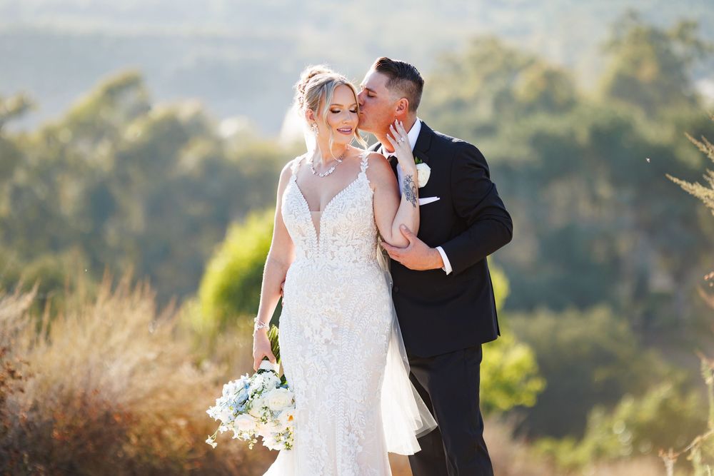 A couple shares an intimate moment in formal wedding attire against a sunlit natural backdrop with dried grasses.