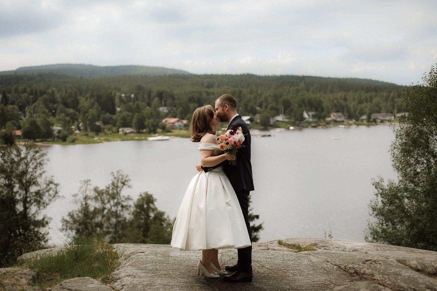 Ett brudpar som står på ett berg med vatten i bakgrunden. De är på väg att kyssa varandra.