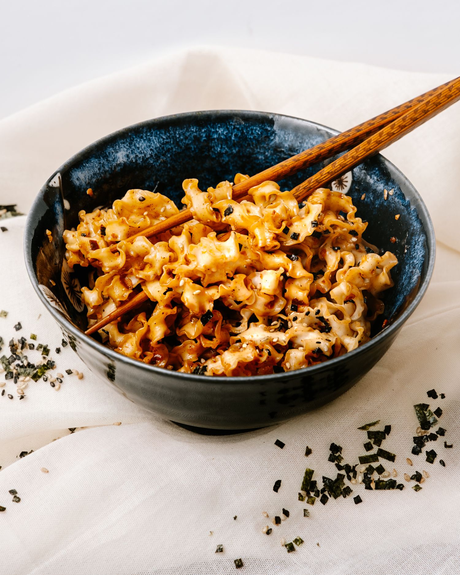 Mac and cheese topped with black sesame seeds served in dark ceramic bowl with wooden chopsticks.