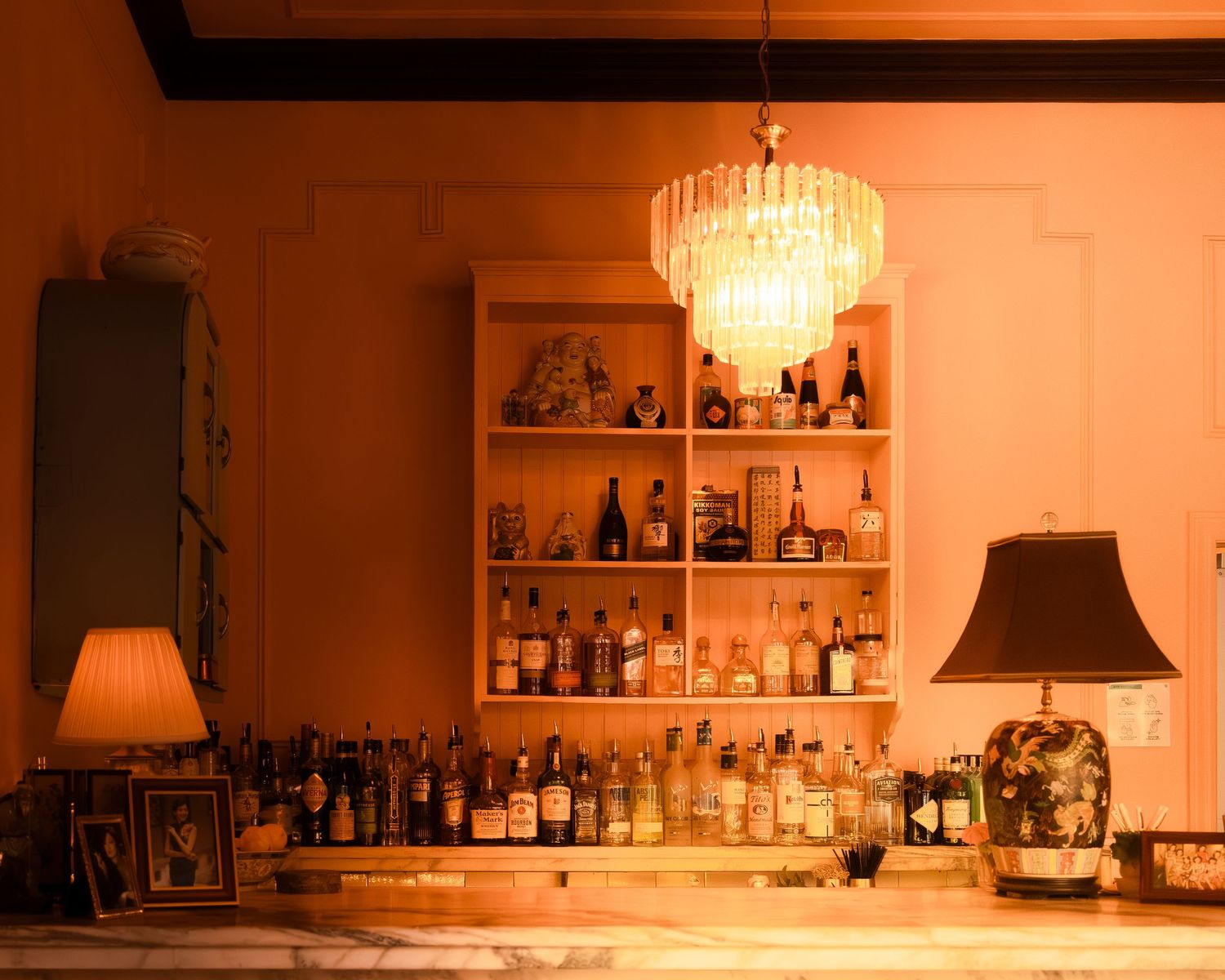 Vintage-style bar interior with crystal chandelier and shelves of liquor bottles in warm lighting.