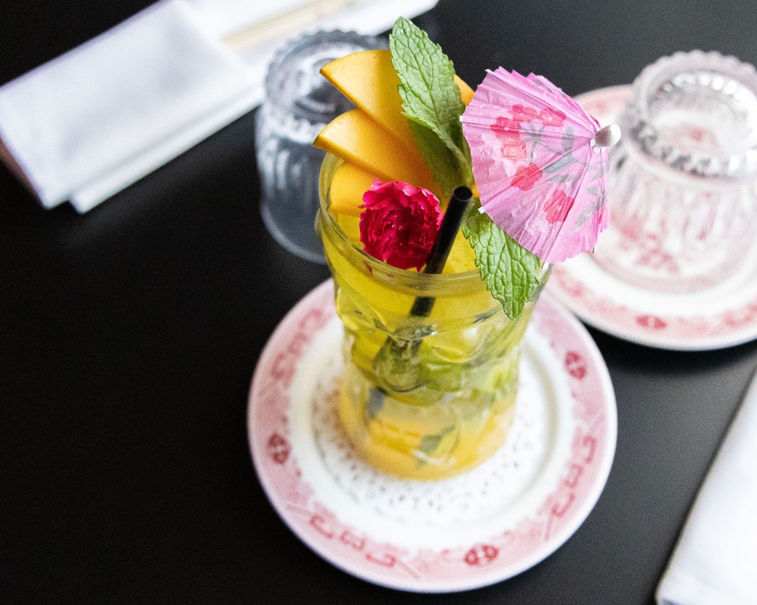 Tropical cocktail garnished with mango, mint, and pink umbrella on floral patterned saucer.