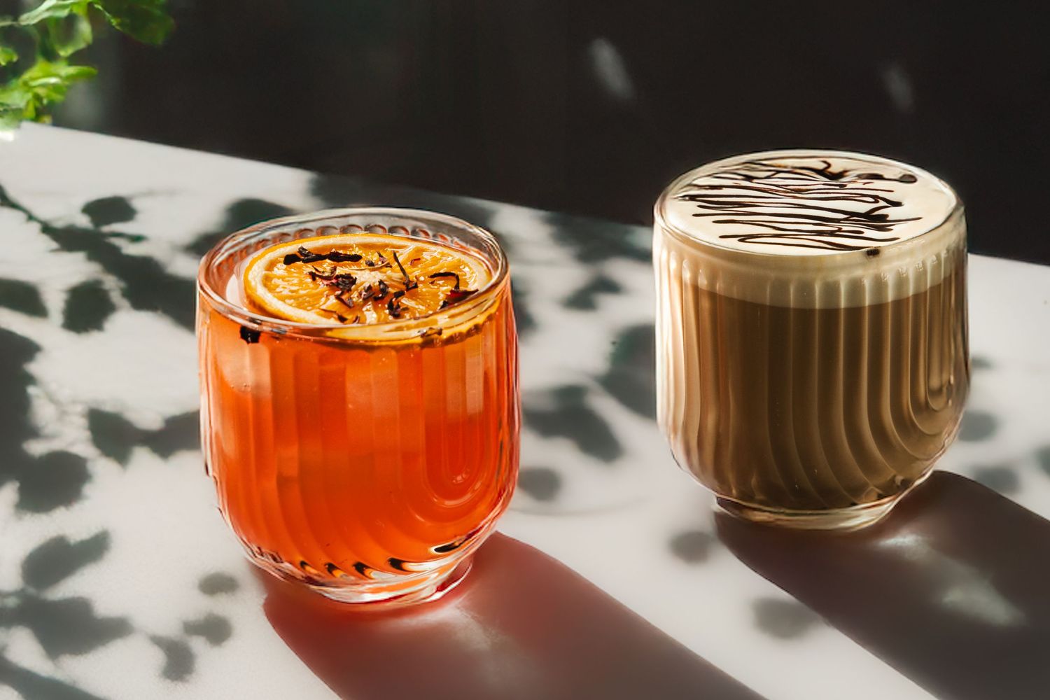 Orange cocktail and chocolate-colored beverage in glass cups casting dramatic shadows in natural light.