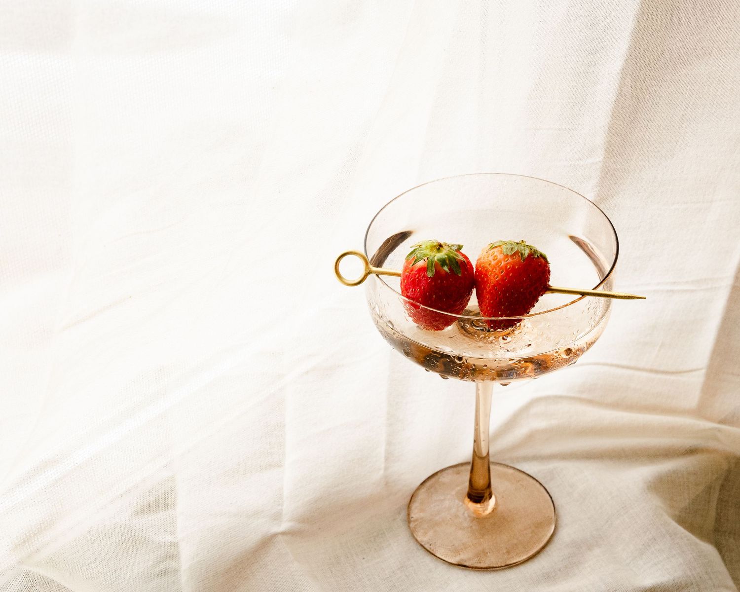 Elegant cocktail glass containing strawberries on white fabric background.