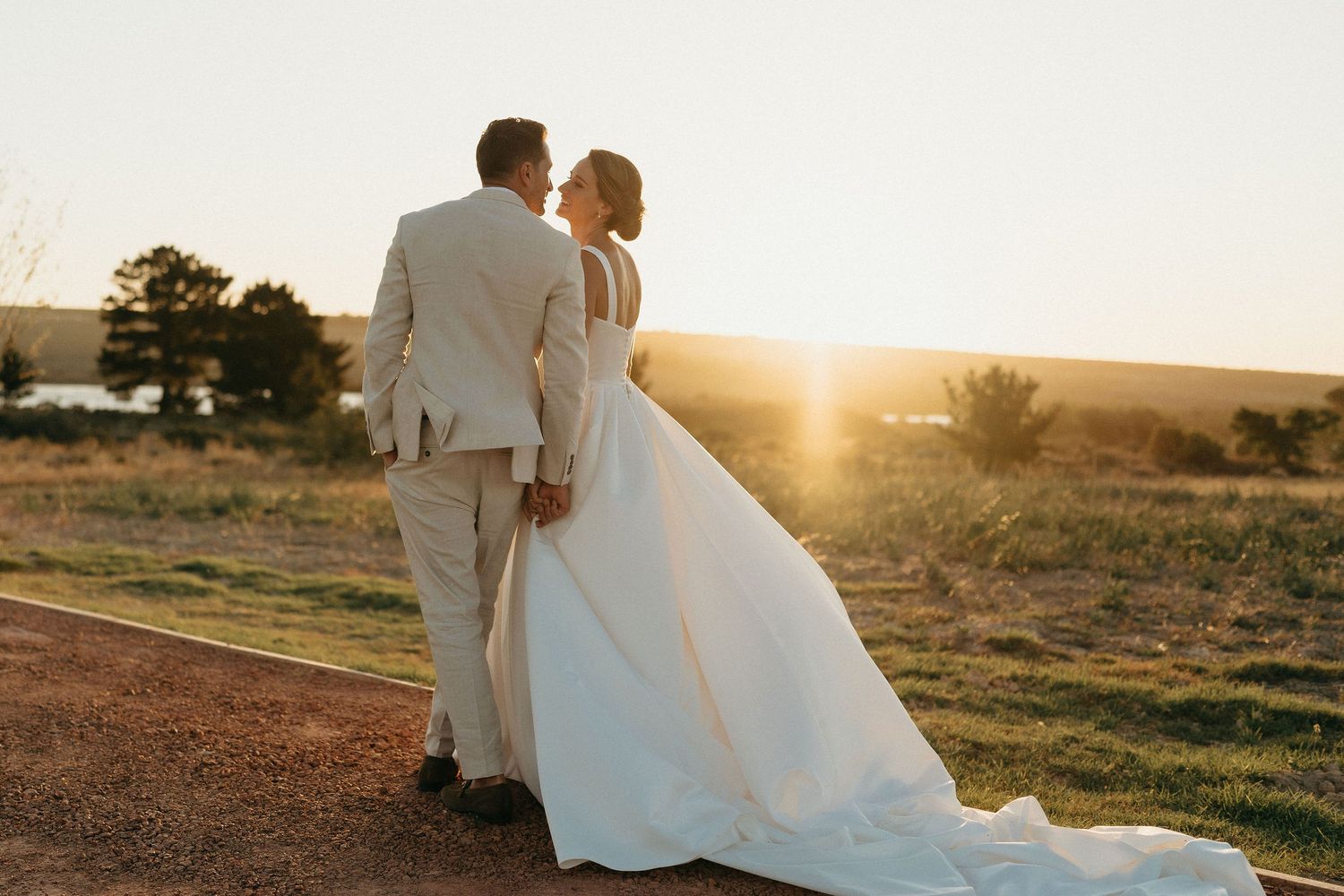 A couple in wedding attire stands together on a dirt path during golden sunset in a rural landscape.