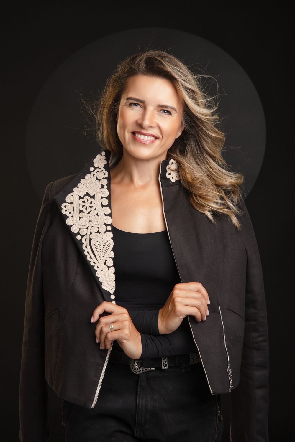 Fashion portrait of someone in a black blazer with ornate white lace detail against dark background.
