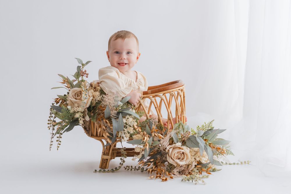 A baby in neutral tones sits on a white floor surrounded by soft beige floral arrangements and dried flowers.