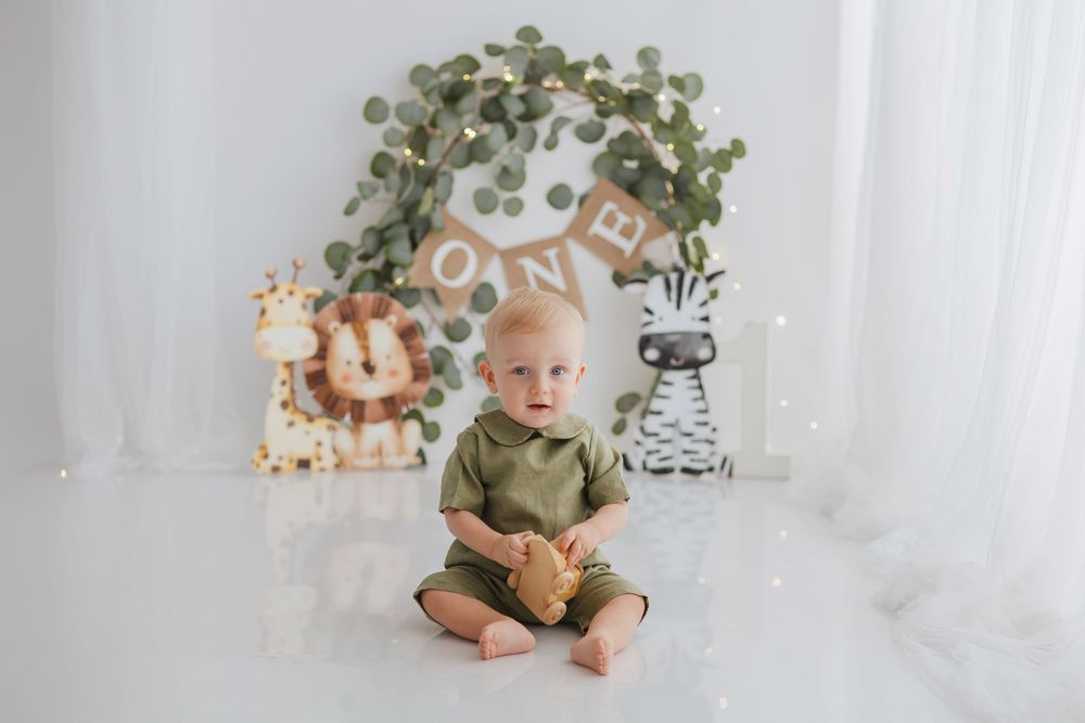 A safari-themed first birthday party setup with greenery arch, jungle decorations and light neutral backdrop.