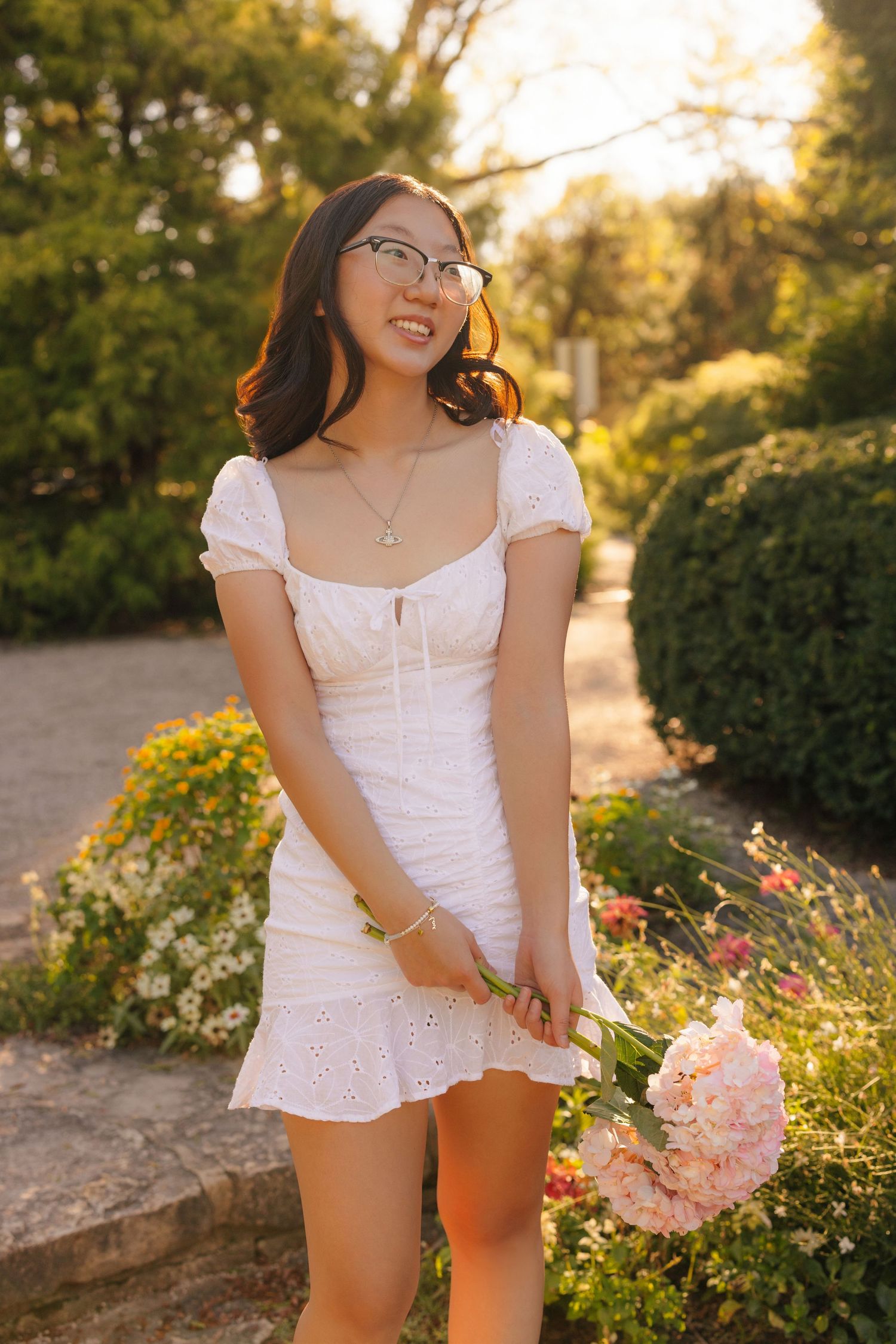 Person in white eyelet dress holding pink flowers in a sunny garden setting.