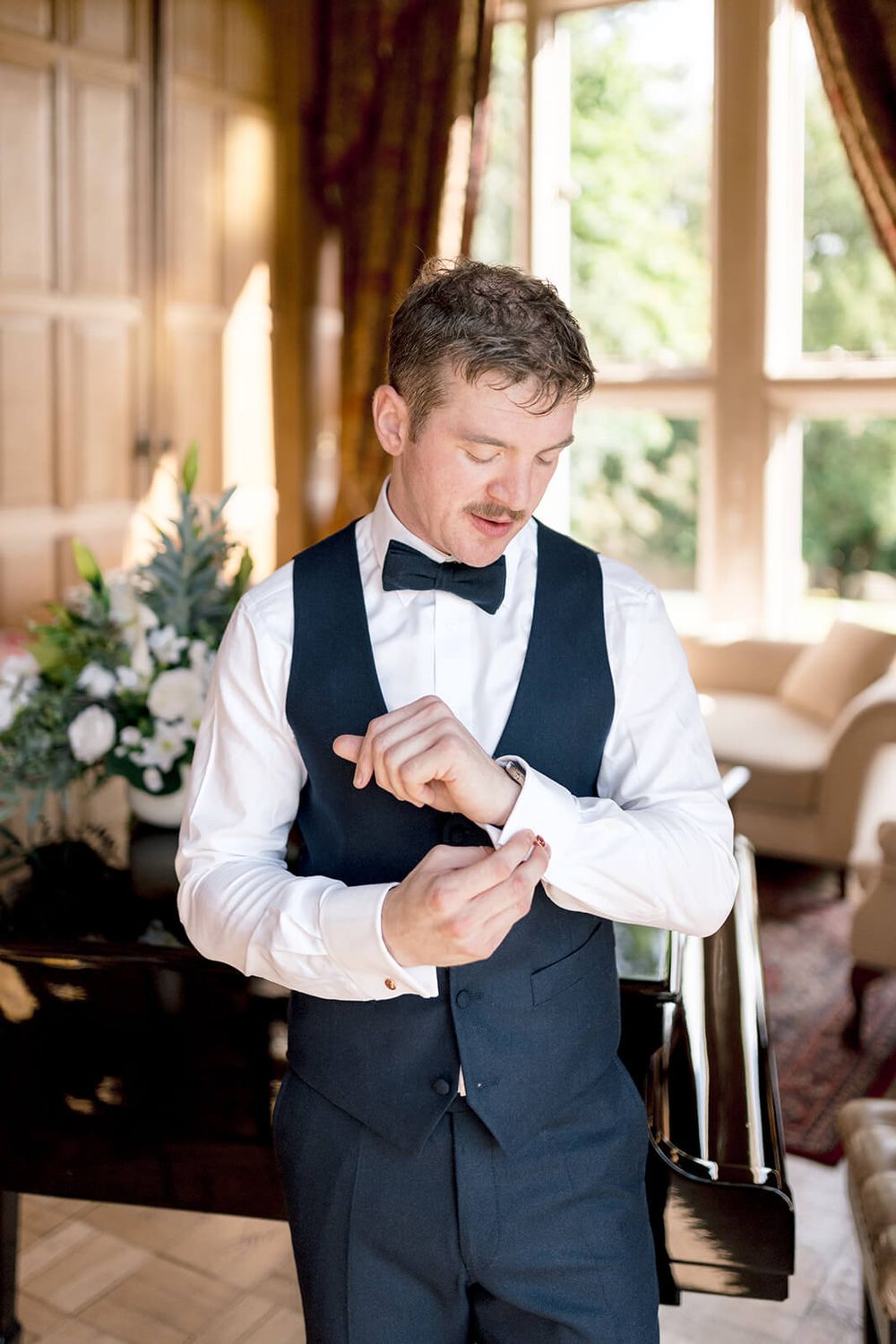 Formally dressed groom adjusts their cuff links while getting ready in an elegant room with natural light captured by Dina Ozola Photography