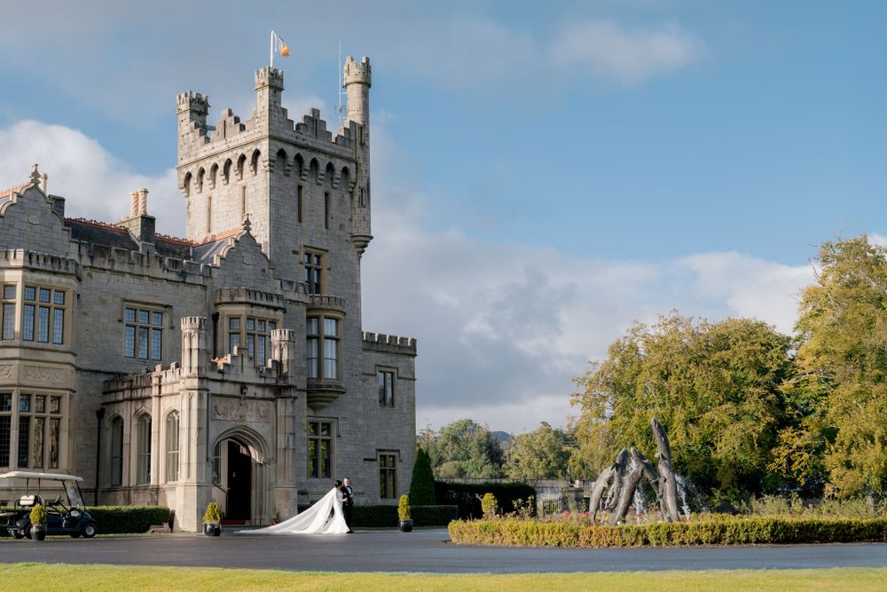 Grand stone castle with Gothic tower and beautiful grounds features a wedding celebration with flowing white dress in sunlight.