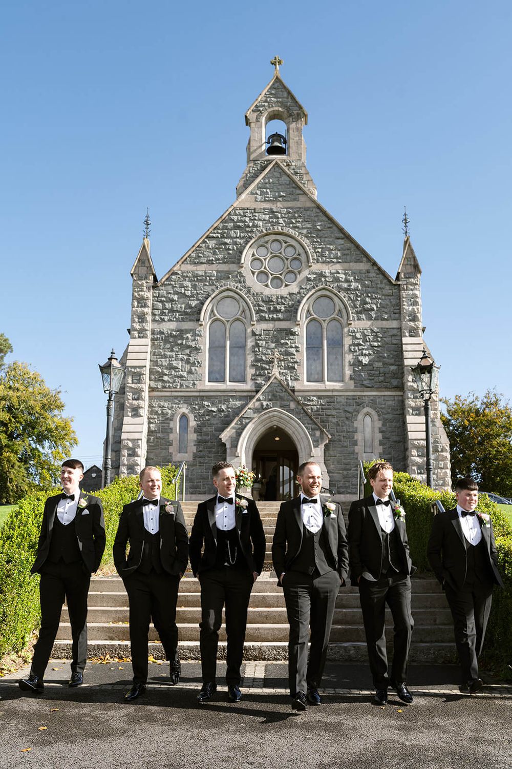 Group of groomsmen in black tuxedos stand on church steps with Gothic stone architecture.