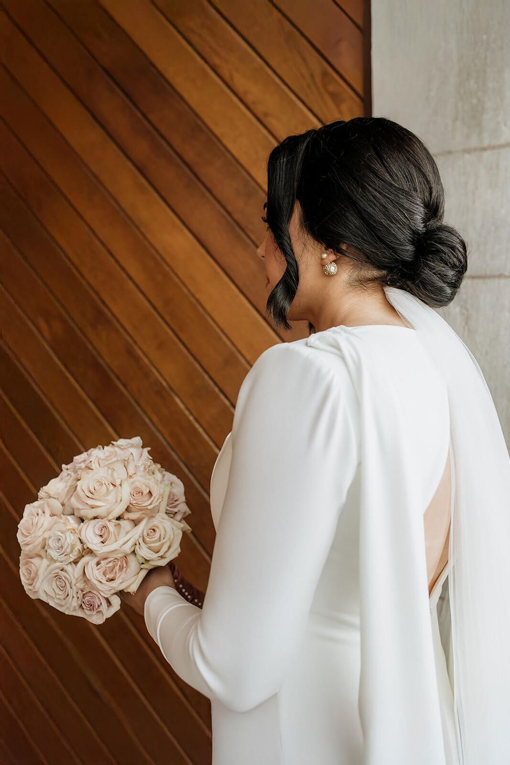 Elegant bride in white long-sleeved dress holds blush rose bouquet against wooden door.