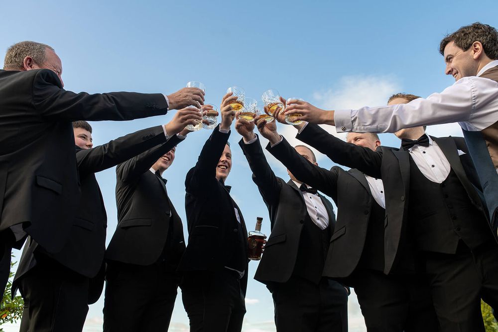 Groomsmen in suits raise their glasses in a celebratory toast against blue sky, captured by Dina Ozola Photography.