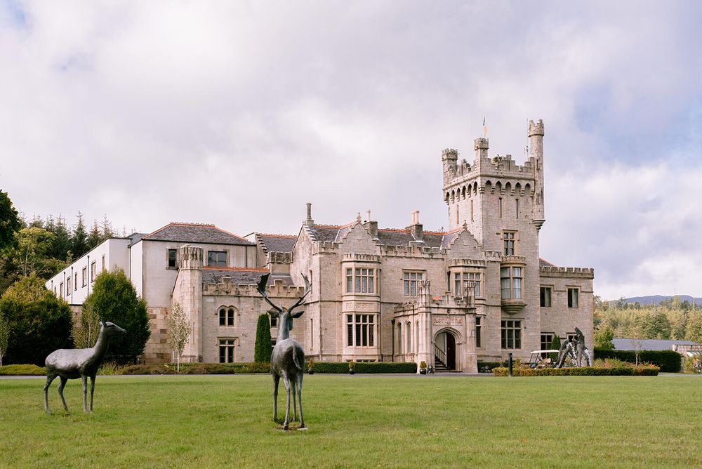 Majestic stone castle with turrets stands on green lawn with decorative deer sculptures. Lough Eske castle, ireland.