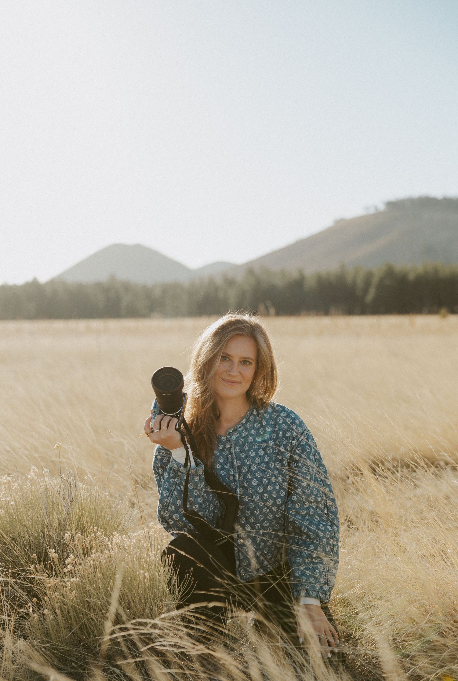 Photographer with camera kneels in tall golden grass at sunset with mountains and forest in background.