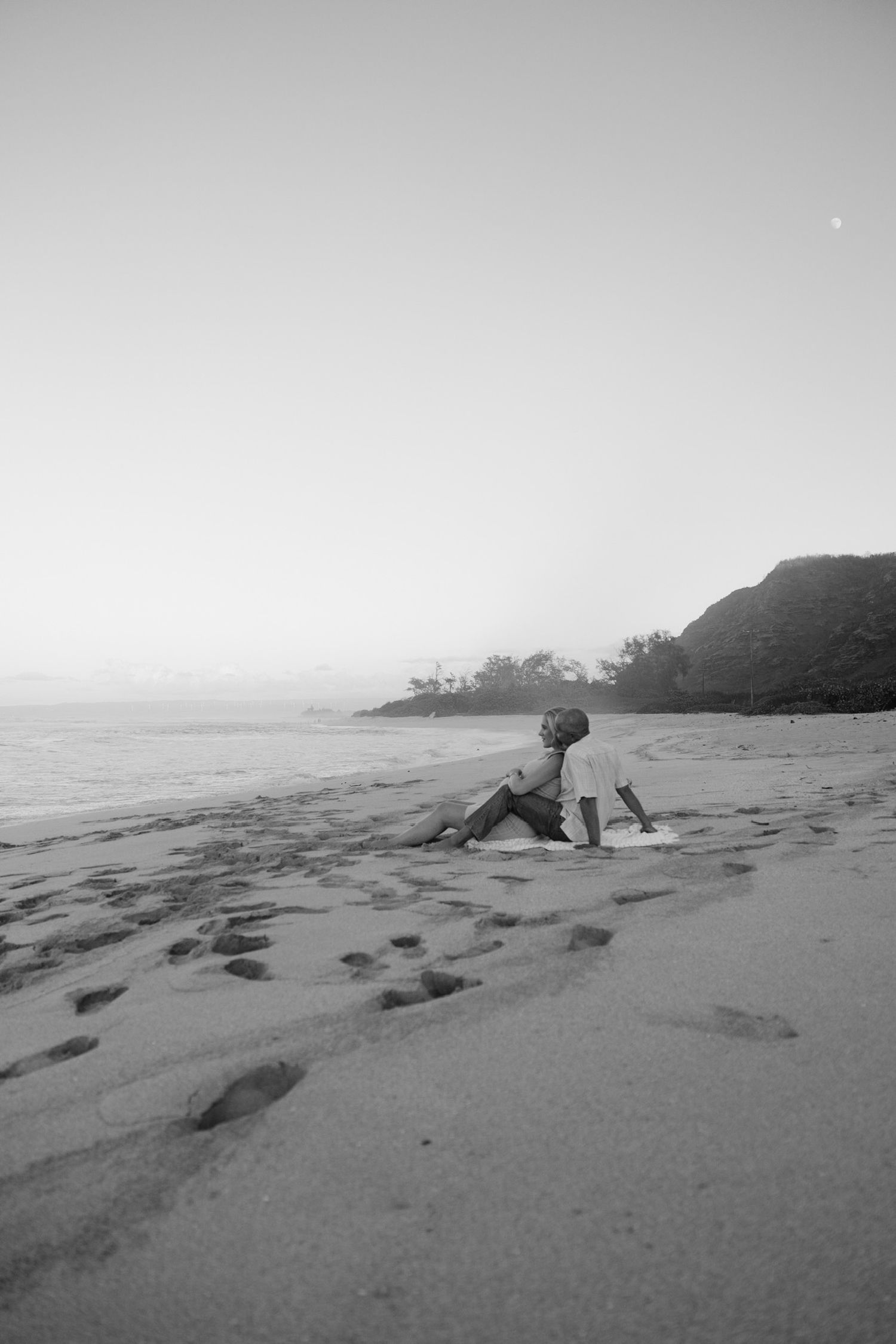 A person sits on a sandy beach at sunset with mountains and ocean in the background, captured in black and white.
