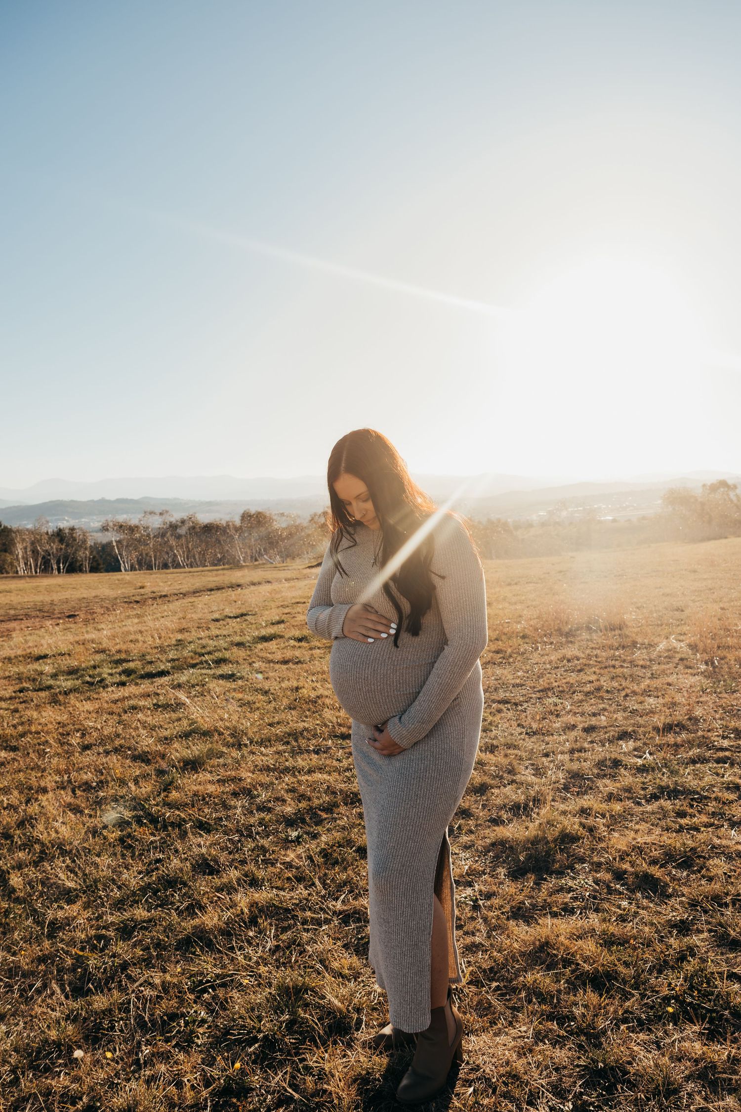 Expectant mother in flowing dress holds belly in sunlit rustic field setting.