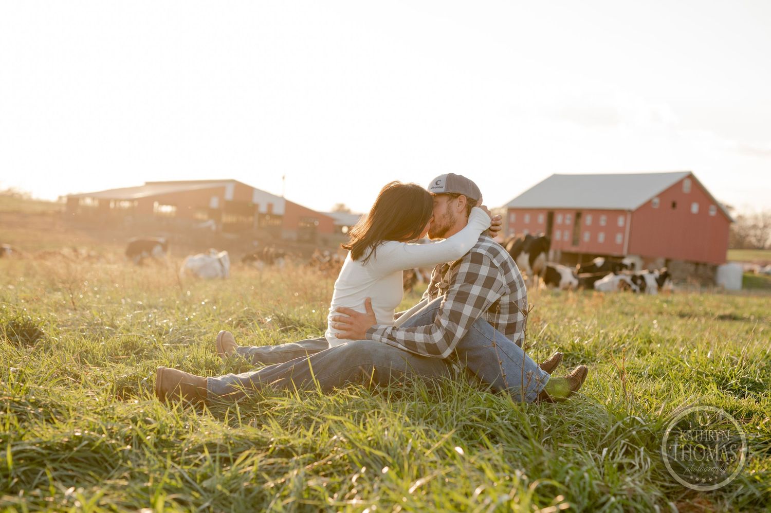 A couple relaxes together on grassy farmland during sunset with a red barn in the distance.