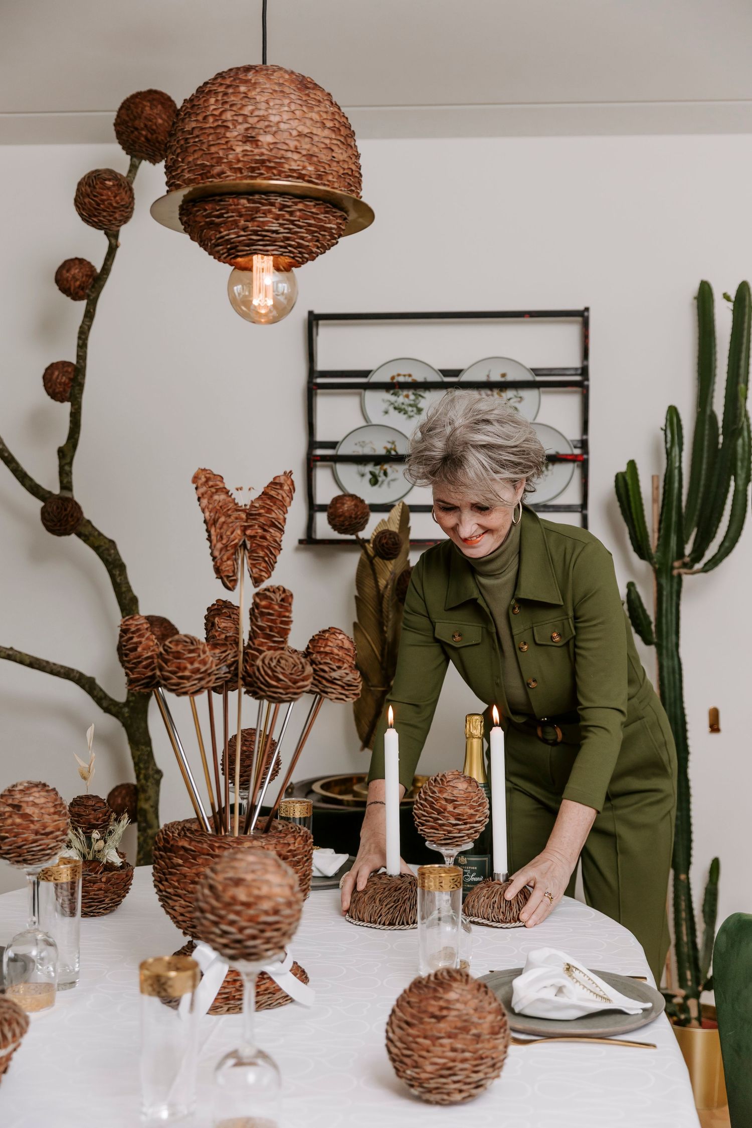 Kvinde i olivengrøn kjole, der dækker bord med flettede dekorationer og kaktusplanter i moderne interiør. Brandingfotograf Fyn