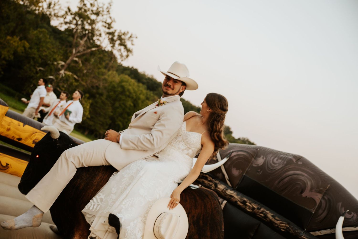 Bride and groom in western attire ride a horse together at outdoor wedding reception.