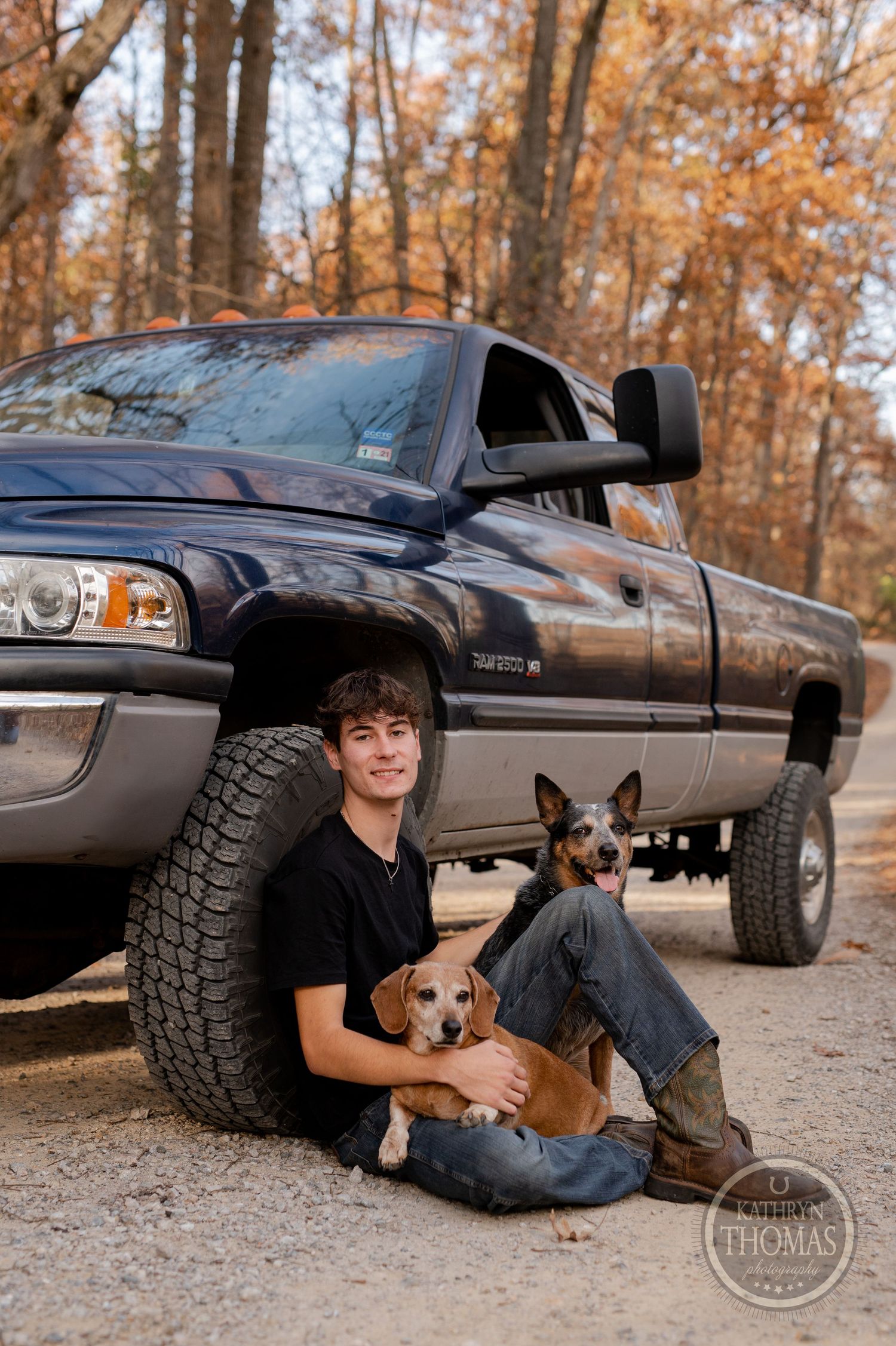 Ram pickup truck with two small dogs and someone sitting by tire in autumn forest setting.