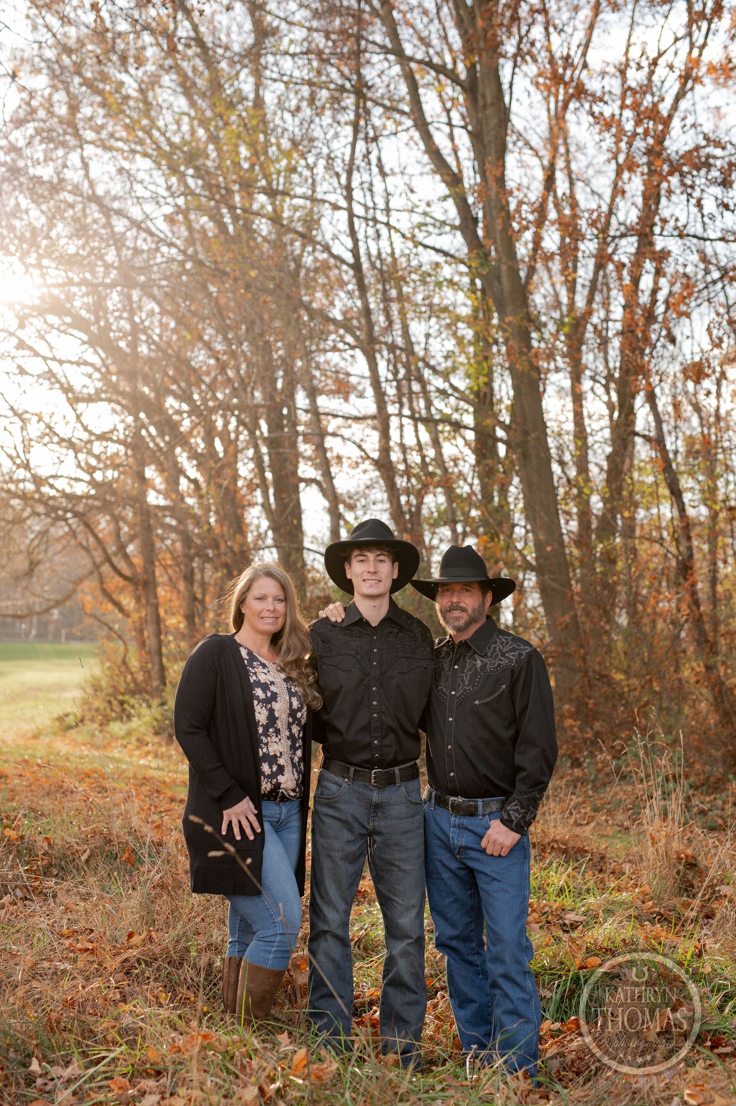 Family portrait of three people in western attire against autumn forest backdrop.