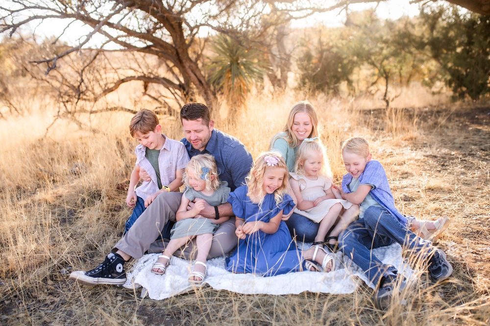 Father sitting on blanket in golden field surrounded by six happy children in blue outfits.