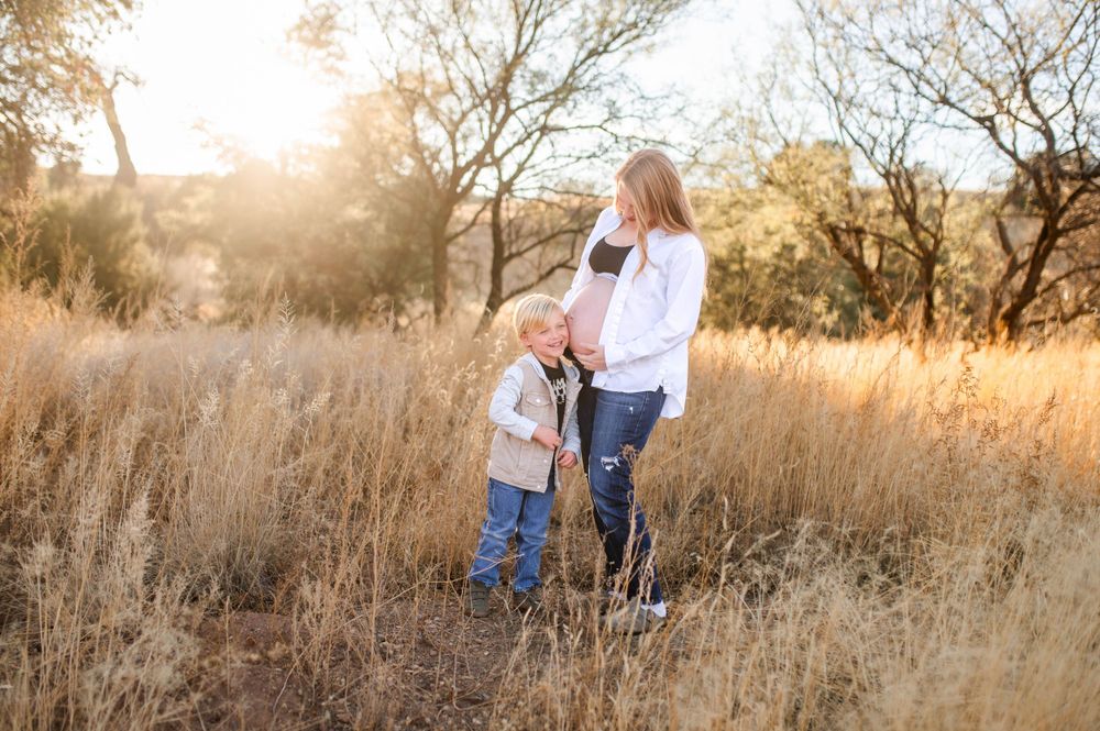 Pregnant mother with young toddler son standing in golden field of tall grass during sunset.