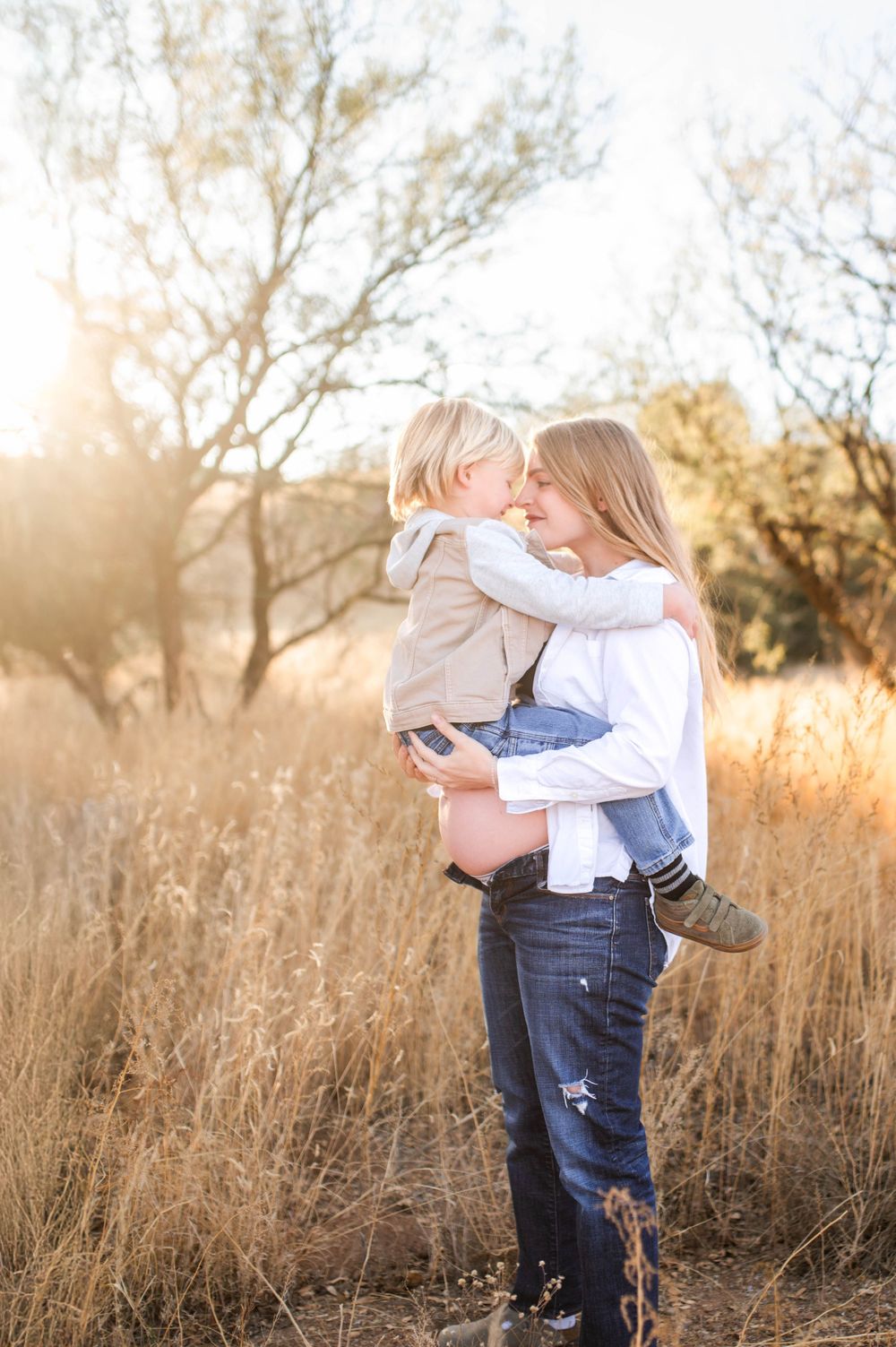 Pregnant mother holding and kissing her young blonde toddler son in a sun-drenched golden field.