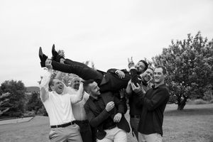 Group of groomsmen lifting groom in black and white outdoor wedding celebration photo.